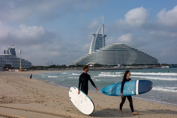 Two people in wetsuits carry surfboards on Jumeirah Beach with the Burj al Arab hotel in the background.
